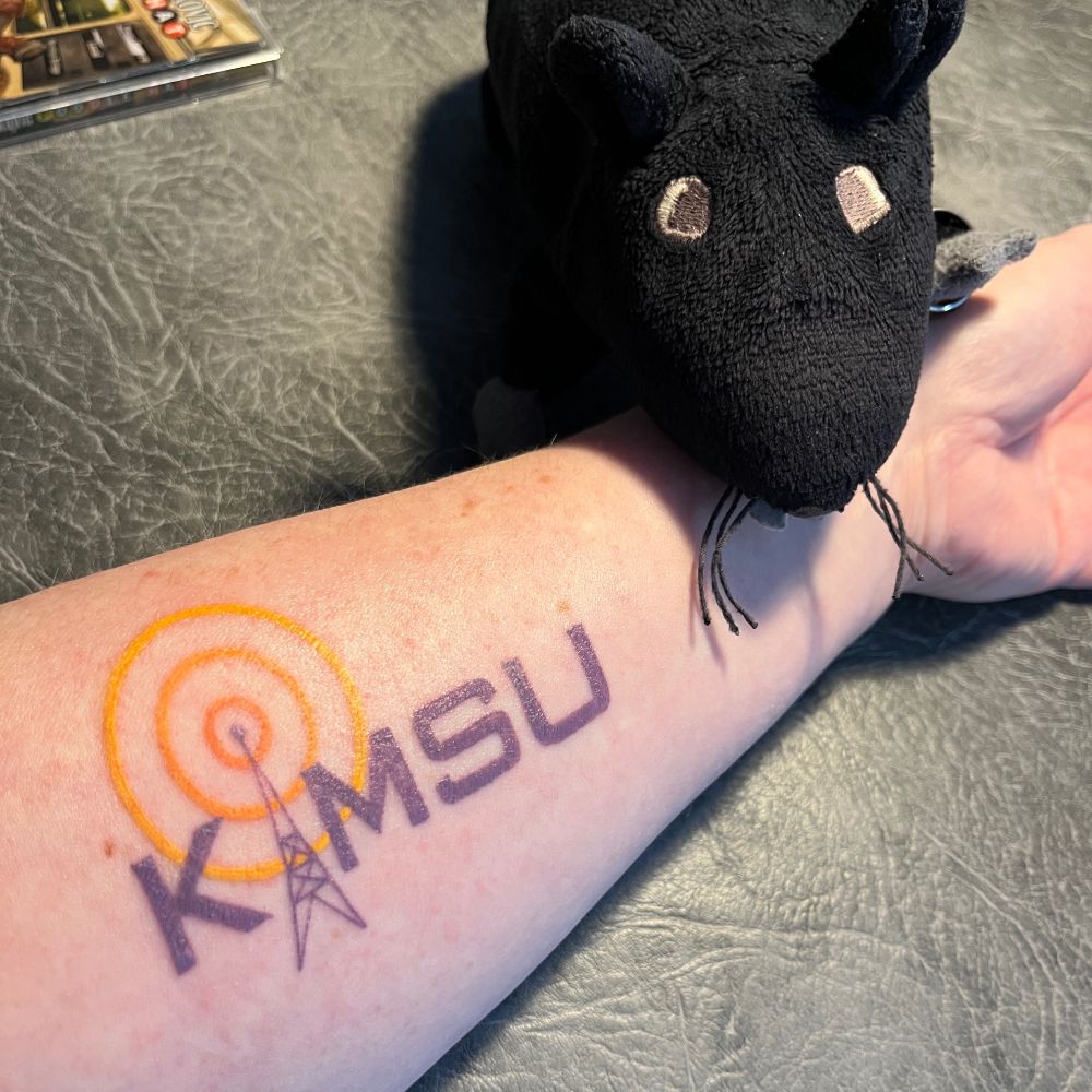 A snap of RM3's left forearm w a KMSU logo tattoo. Zeke balances on her wrist, looking like he's grabbed and dragged the bodypart onto the table.